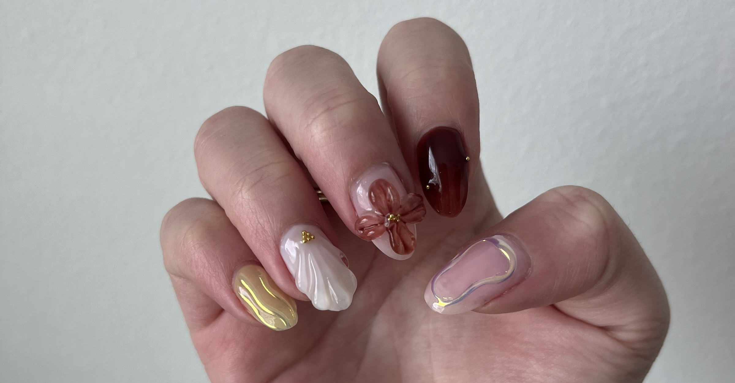 A hand with an almond-shaped set: butter-yellow chrome, white marble, a hand-painted terracotta bow, oxblood with gold beads and an iridescent swirl.