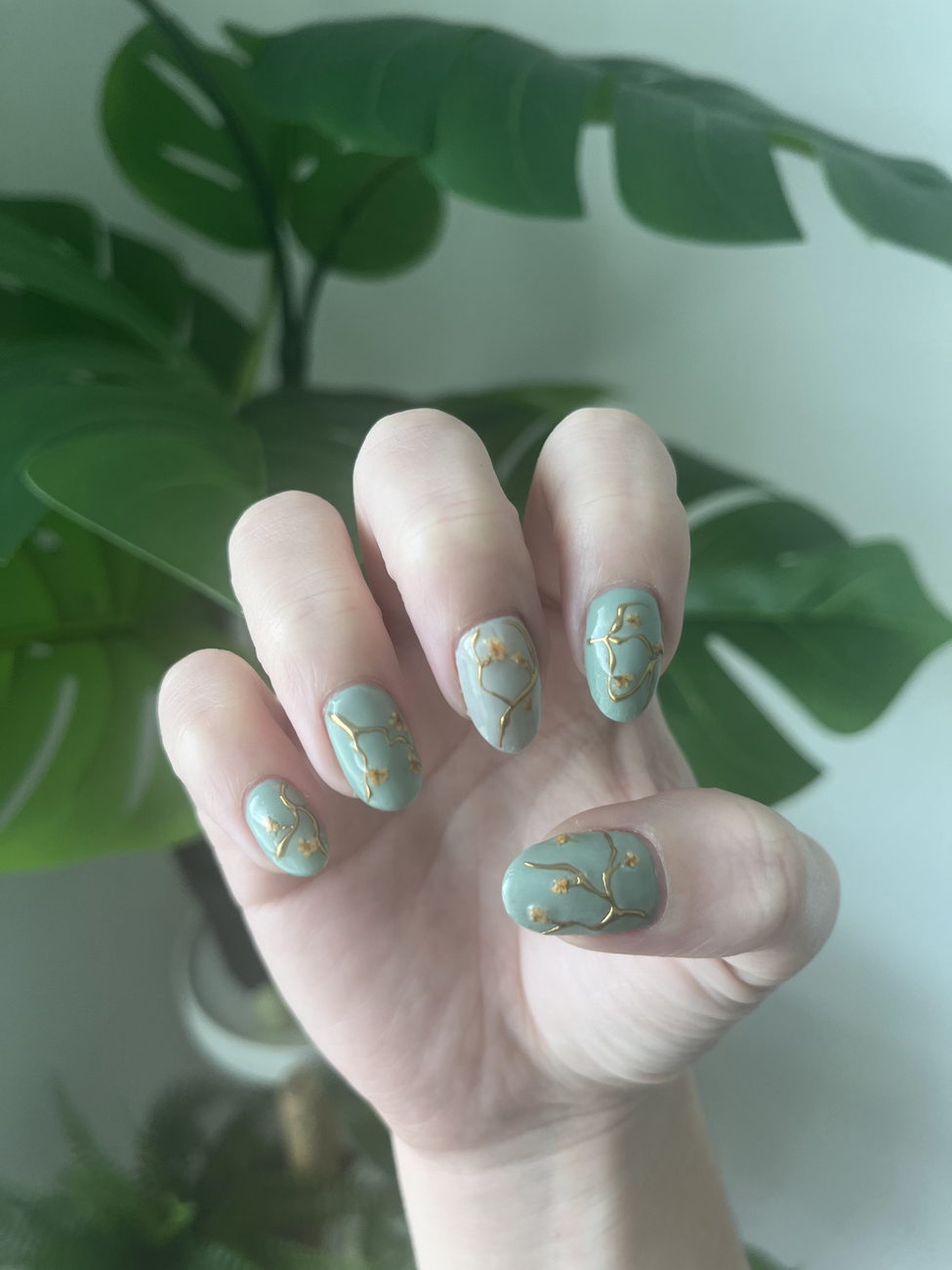 Short oval nails in soft sage green, each with a fine gold branch and small blossoms, held in front of a monstera plant.