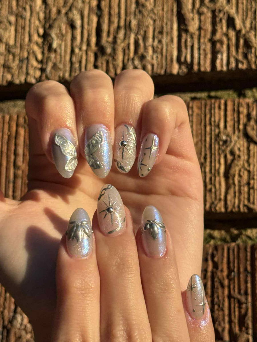 Pearlescent almond nails carrying silver butterfly, flower and starburst charms, photographed against a weathered wood wall in low sun.