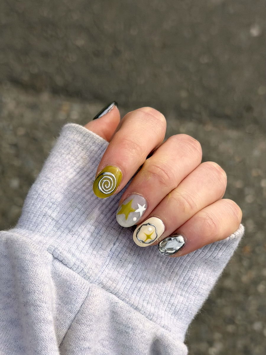 A hand in a gray sweatshirt: short round nails in olive with a silver spiral, gray with painted stars, cream with a pearl, and one in mirror chrome.