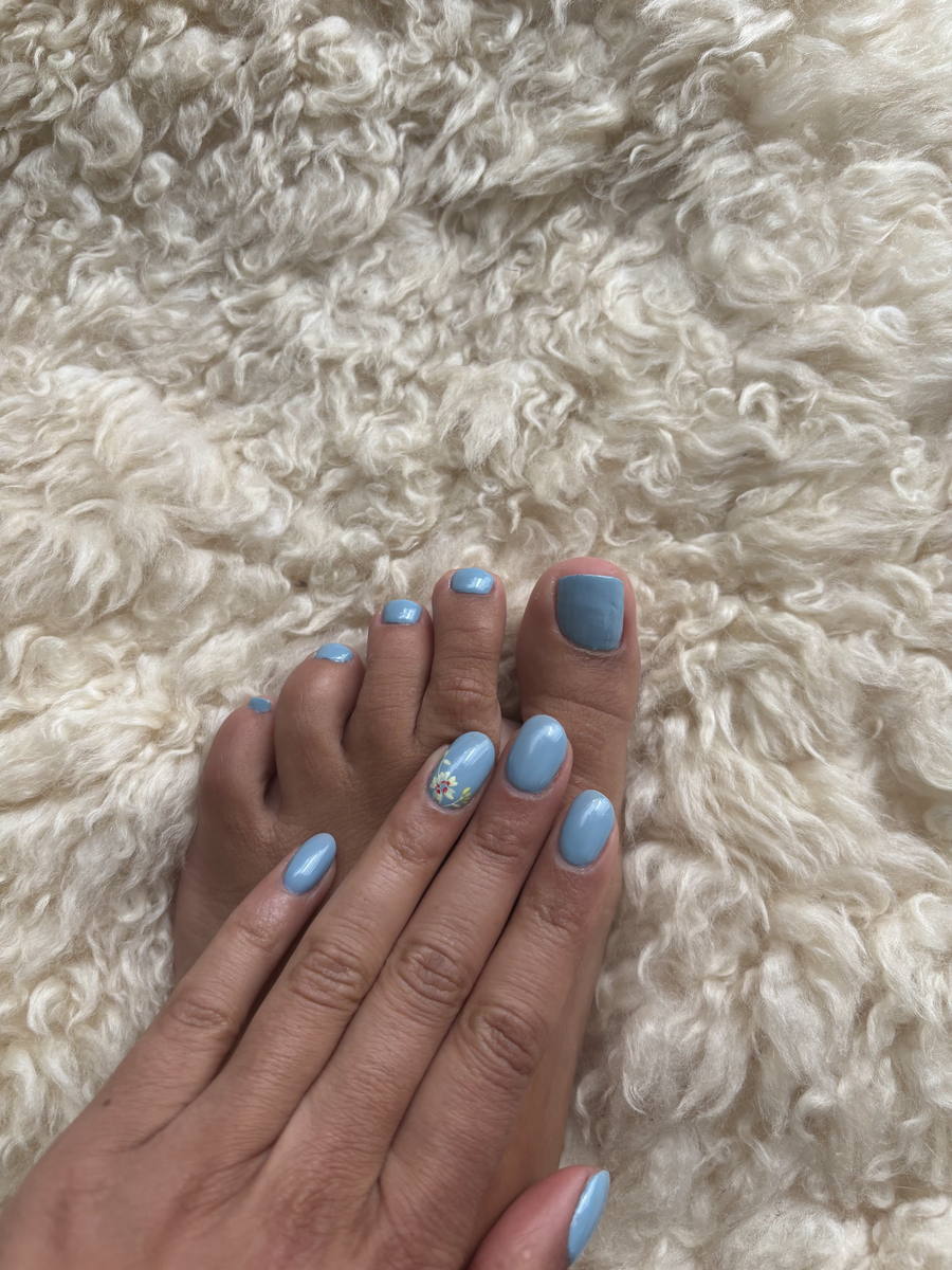 A foot and a hand resting on a cream sheepskin rug, every nail in sky blue, one finger carrying a small painted flower.