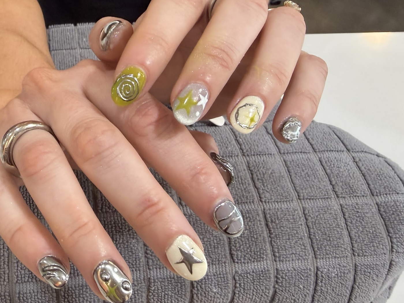 A guest's hands resting on a gray towel at the manicure table, wearing chrome, star and lemon designs.